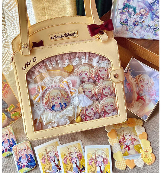 Cream ita bag decorated with anime pins/badges and a plush insert, styled on a desk with matching character cards and merch.